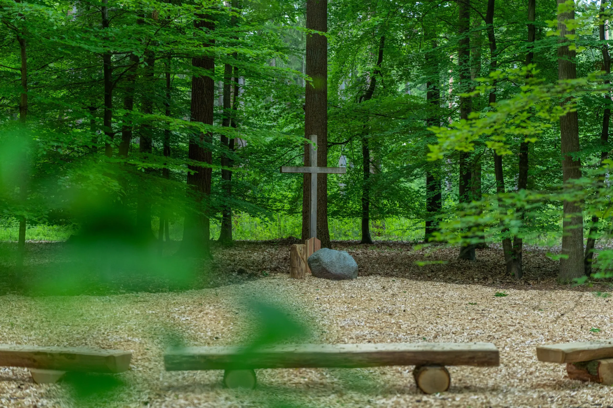 Andachtsplatz im Wald mit einem schlichten Holzkreuz, einem Stein und Sitzbänken zwischen grünen Bäumen.